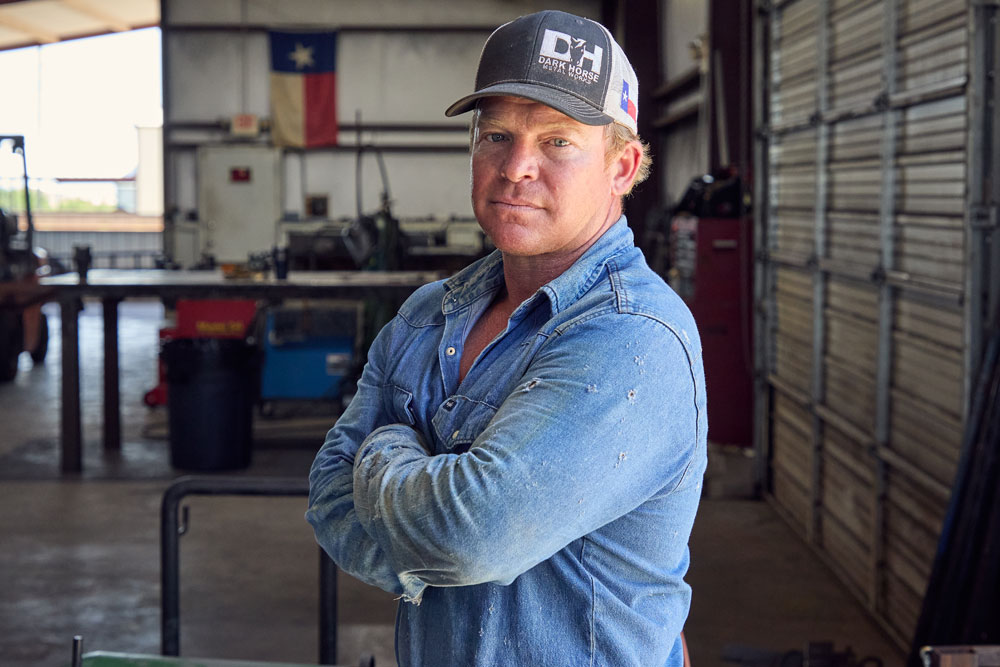 Un hombre, Wes Stevenson, con una camisa vaquera desgastada y una gorra de béisbol, está de pie con los brazos cruzados en un taller de metal, mirando con confianza a la cámara; la bandera de Texas cuelga de la pared detrás de él.